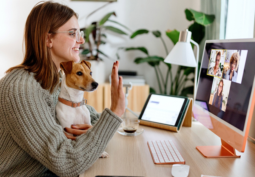 Photo of a female student at home sitting at her desk with a dog in her lap. She is waving toward her laptop, where three people are shown on the screen.