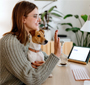 Photo of a female student at home sitting at her desk with a dog in her lap. She is waving toward her laptop, where three people are shown on the screen.