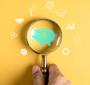 A hand holds a magnifying glass over an image of a green piggy bank. Smaller images are depicted in a circle around the magnifying glass that include a graduation cap, a pie chart, two hands shaking and a target with an arrow in the center.