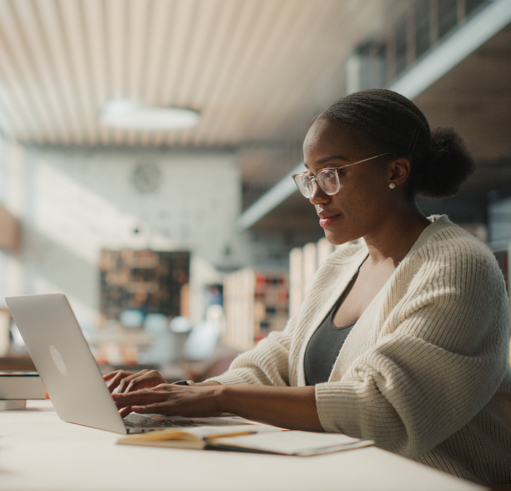 Focused student wearing glasses works on a laptop at a library table, with bookshelves and natural light in the background.