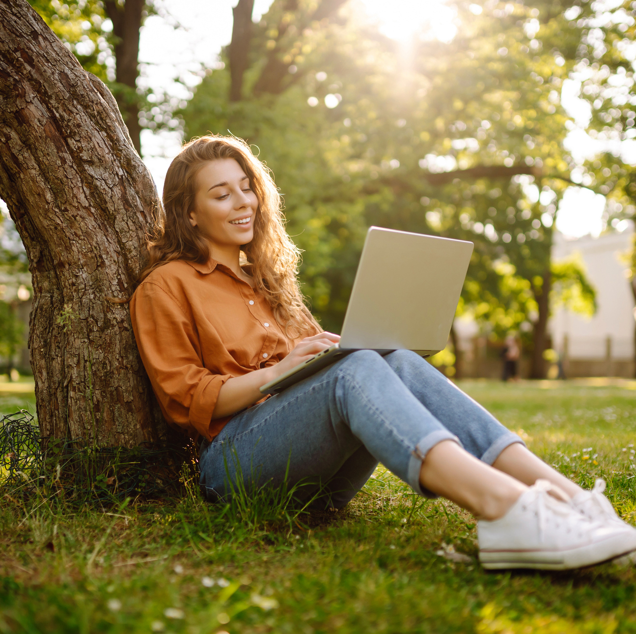 An adult female student sits outside leaning against a tree with a laptop balanced on her knees.