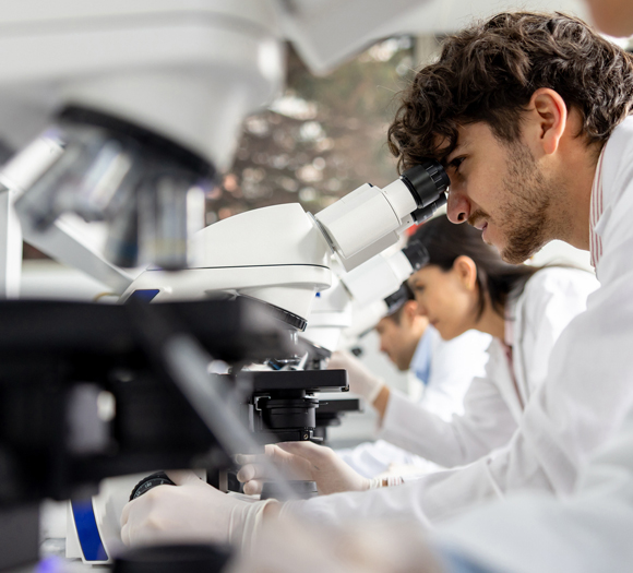 Young scientists in lab coats and gloves focus intently on microscopes in a laboratory. The mood is concentrated and collaborative.