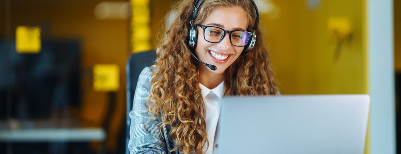 A photo of female student with long hair and glasses wearing headphones as she communicates with someone on her laptop.