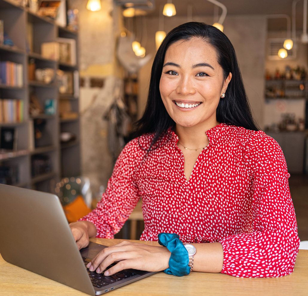 A photo of a female adult student smiling at the camera as she sits at a table in a public space working on a laptop.