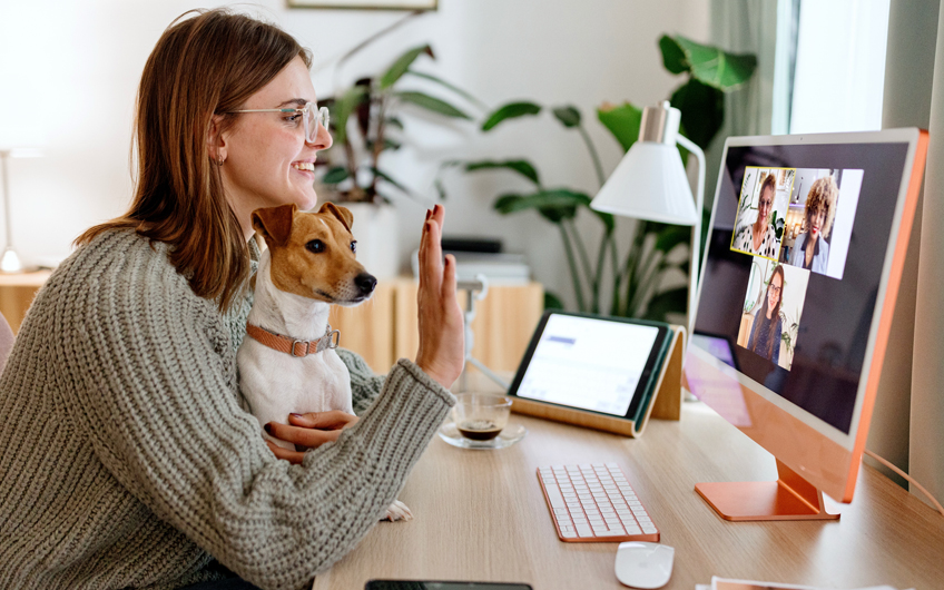 Photo of a female student at home sitting at her desk with a dog in her lap. She is waving toward her laptop, where three people are shown on the screen.