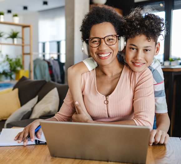 A photo of an African American woman smiling and sitting at a table facing forward while working on a laptop at home. A young smiling boy stands behind the woman with an arm wrapped around her.