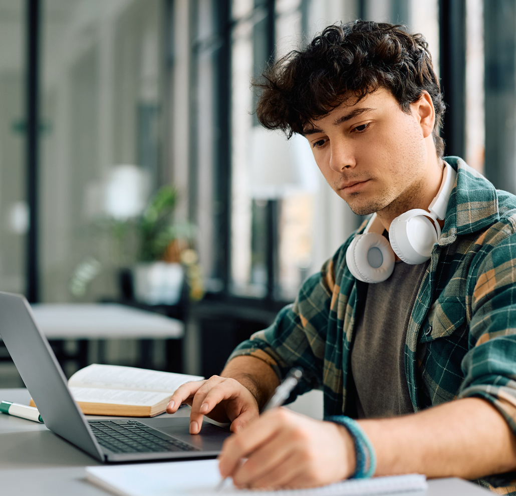 A photo of a young male adult who sits in a public space working on a laptop while writing in a notebook.