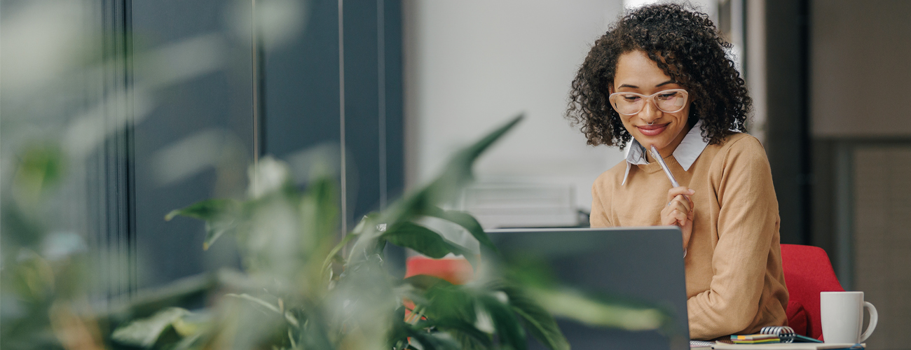 A photo with a blurry plant in the foreground and an in-focus woman working on her laptop in the background. The woman is contemplating what she is reading, and cup of coffee sits on the table next to her computer.