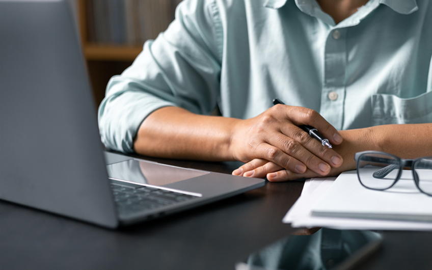 A photo of a person’s arms resting on a table in front of a laptop.