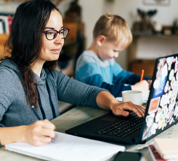 At a dining room table sits a mom doing homework on her laptop while a child sits next to her writing in his notebook.