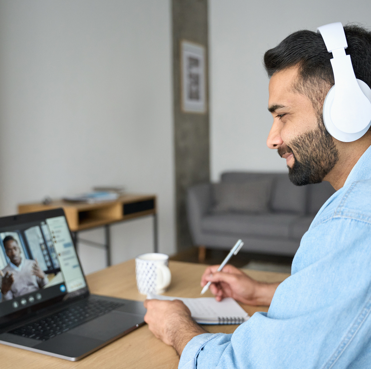 A man with a beard sits at a table with his laptop, taking notes in notebook. On the laptop screen is a person talking.