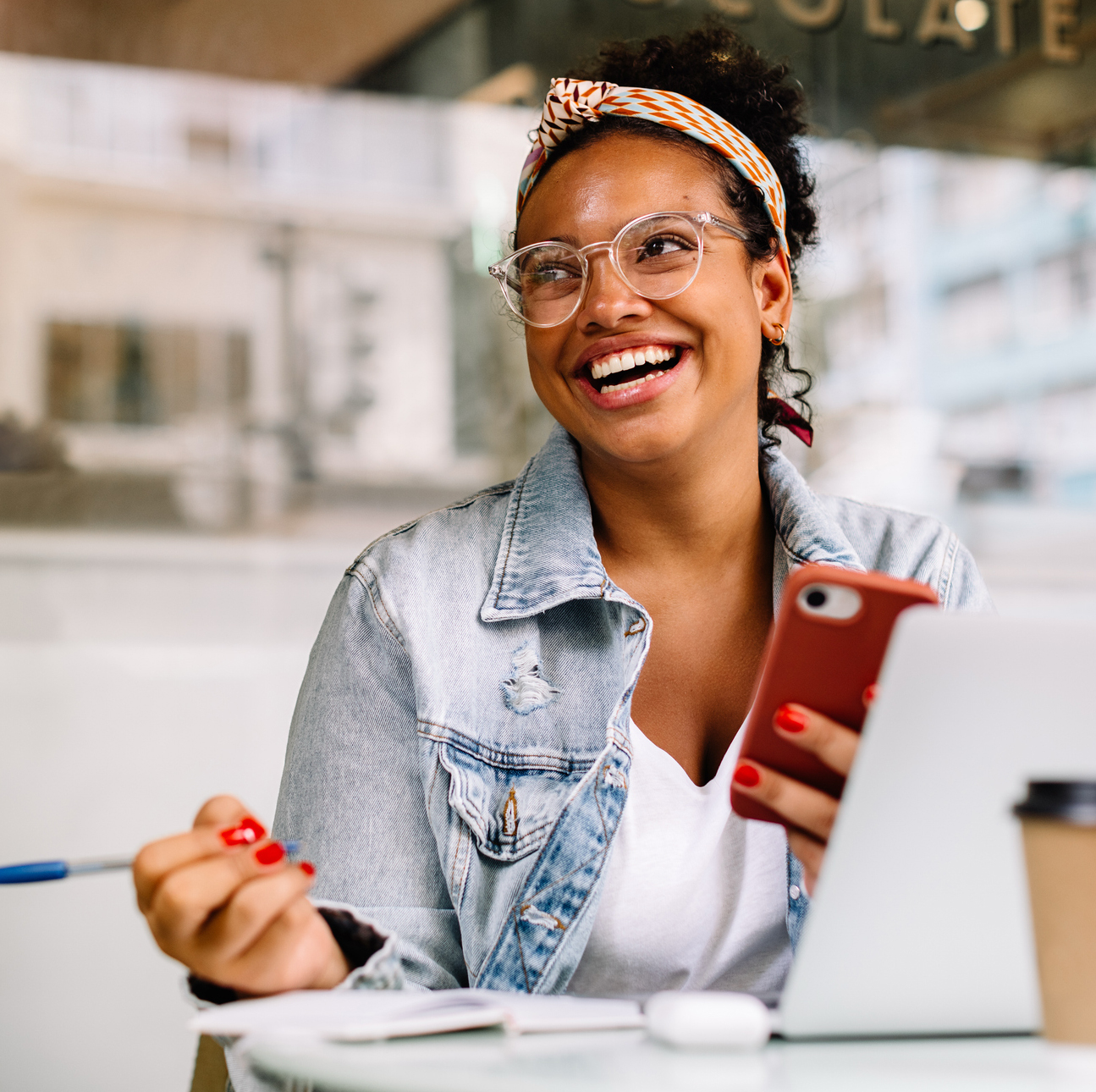 An adult African American woman sits smiling at a table. She holds a pen in her right hand and a cellphone in her left hand. On the table is a laptop and a notebook.