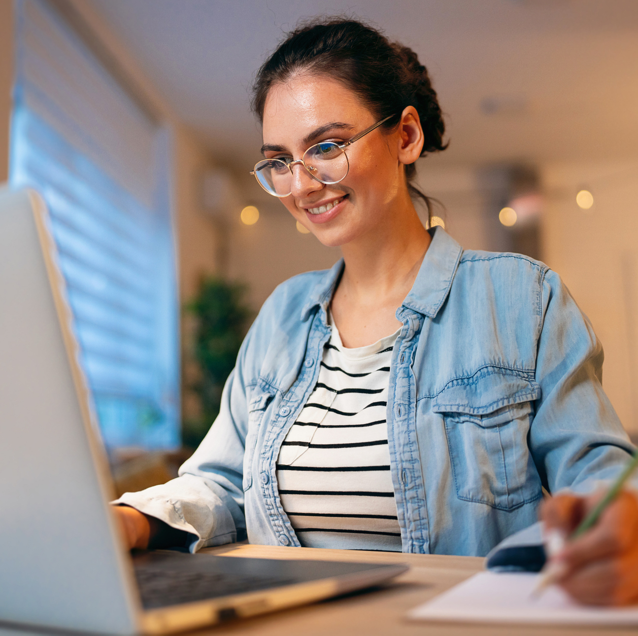 A young woman smiles as she works on her laptop in a home setting.